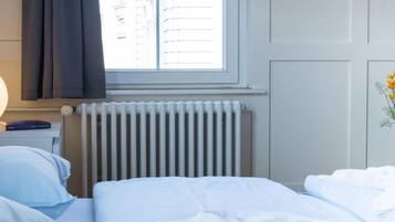 Une chambre d’hôtel avec un lit, une table de chevet avec une lampe, une fenêtre avec des rideaux, un radiateur et un mur orné d’un tableau.