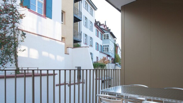 A balcony with a metal table and chairs, a wooden deck, and a white wall with a tree. - Apartment  by Interhome (Basel)