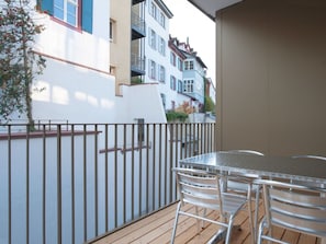 A balcony with a metal table and chairs, a wooden deck, and a white wall with a tree. - BS Hodler II - Marktplatz HITrental Apartment by Interhome (Basel)