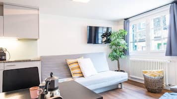 A modern hotel room with a sofa, a dining table, a kettle, and a TV.