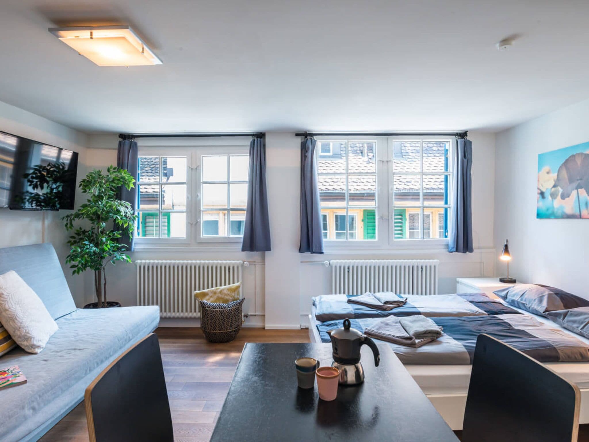 A modern living room with a sofa, a coffee table with mugs, a plant, and a view of buildings through large windows.