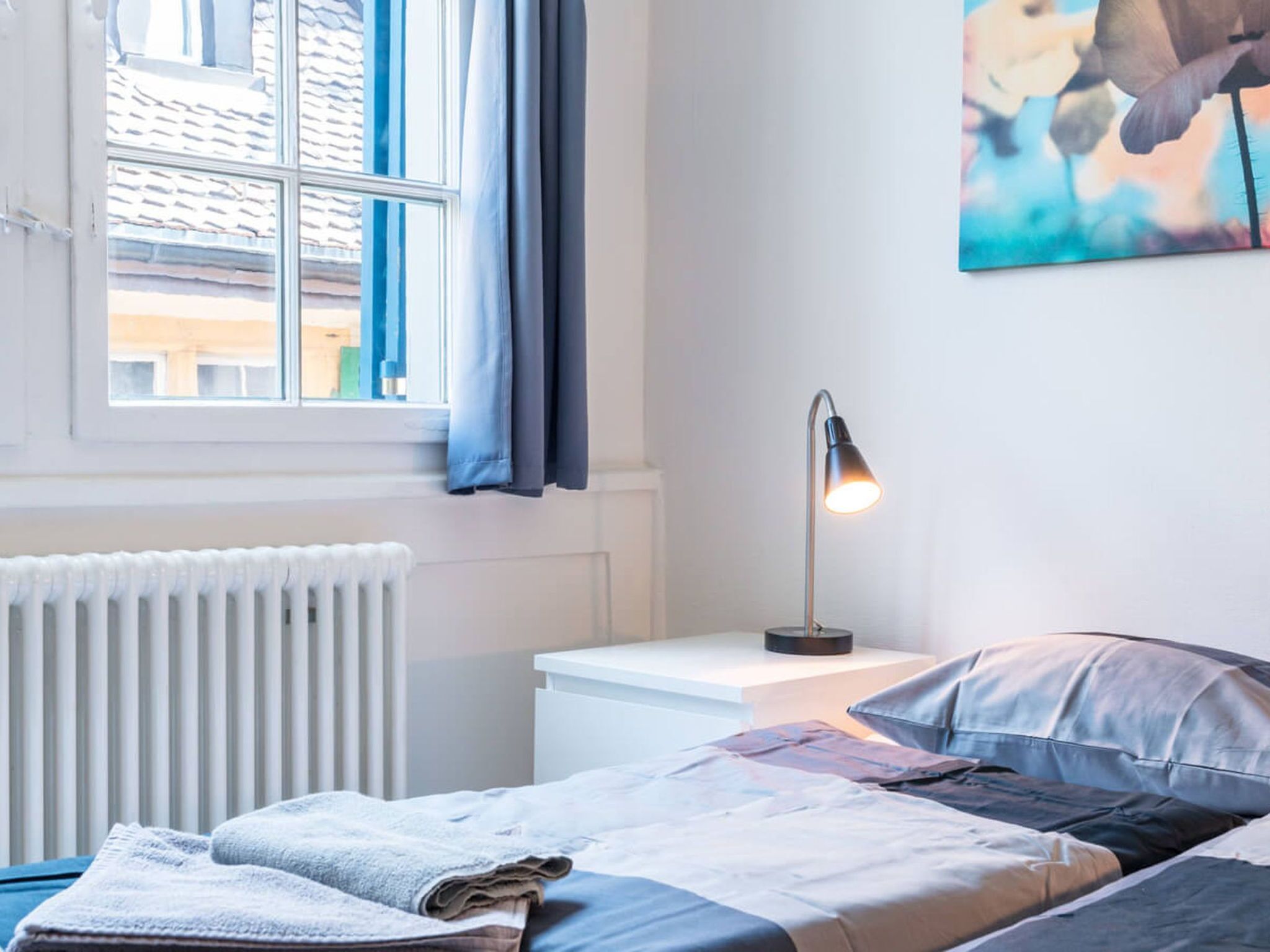 Ein Schlafzimmer mit Bett, Nachttisch mit Lampe, Heizkörper und einem Fenster mit Vorhängen.