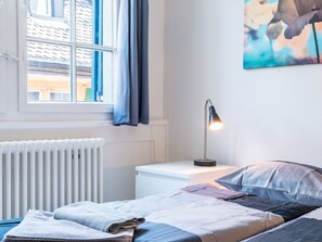 Ein Schlafzimmer mit Bett, Nachttisch mit Lampe, Heizkörper und einem Fenster mit Vorhängen.