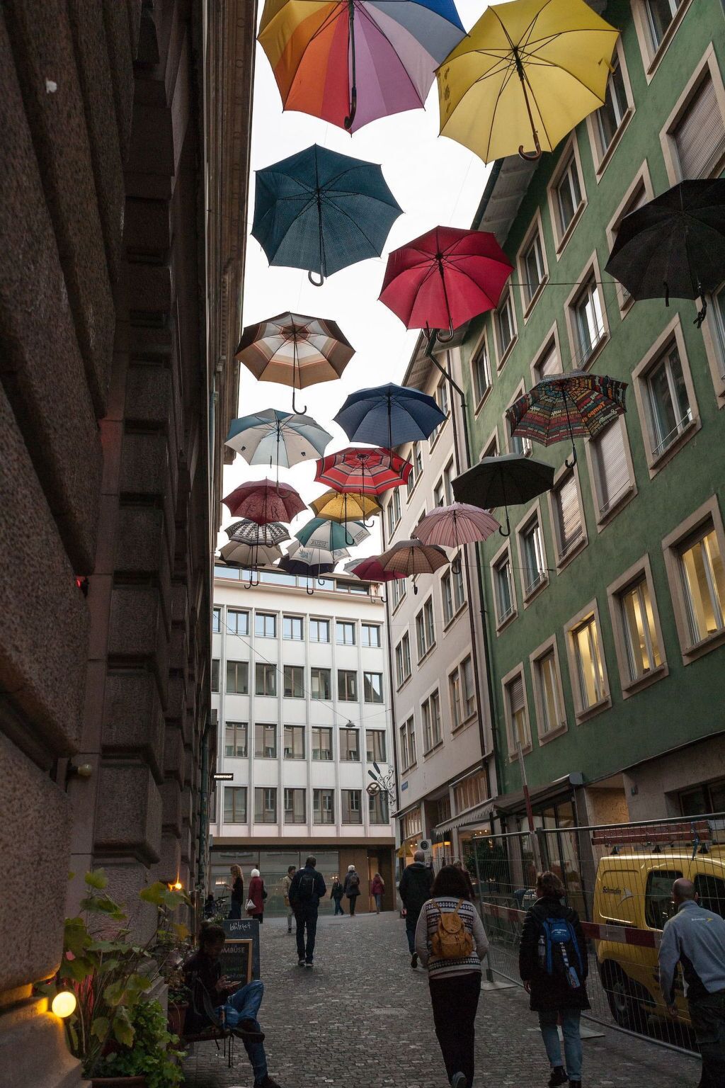 Una calle flanqueada por edificios, con múltiples sombrillas de colores suspendidas sobre ella, y gente caminando.