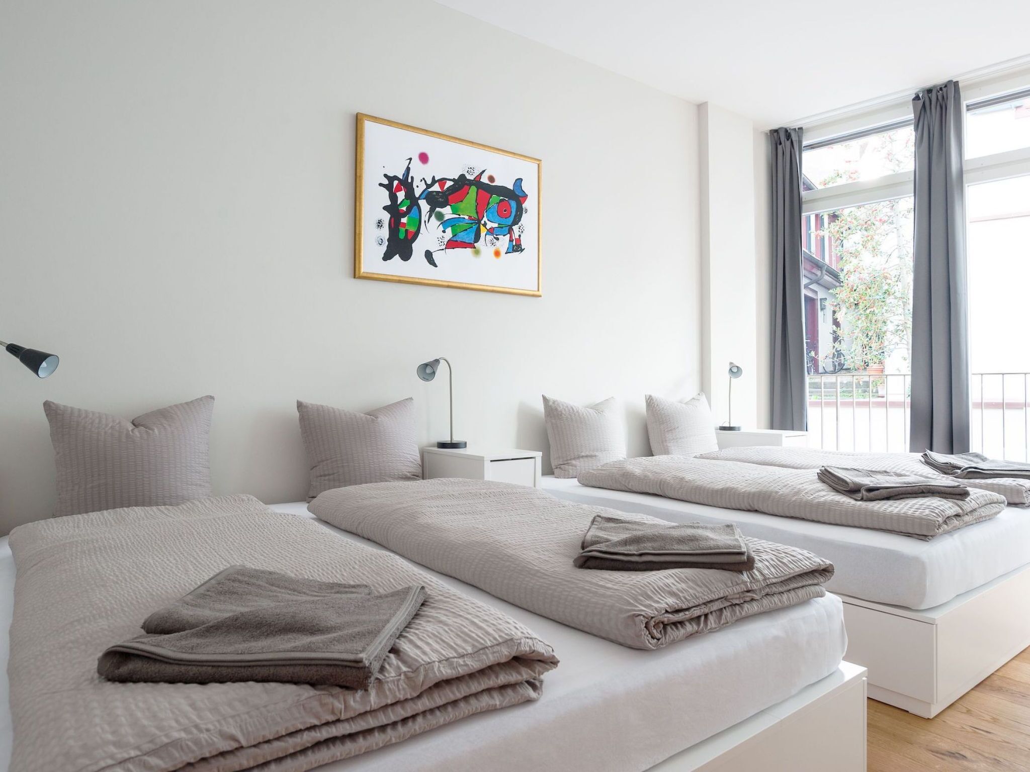 Une chambre d’hôtel moderne avec deux lits, une grande fenêtre avec des rideaux, un tableau au mur et une vue sur l’extérieur.