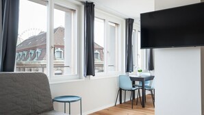 A modern living room with a sofa, a dining table with chairs, a TV mounted on the wall, and large windows with curtains. - Apartment  by Interhome (Basel)