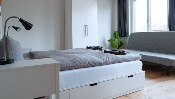A modern bedroom with a bed, a desk, a wardrobe, a plant, and a window with curtains. - Apartment  by Interhome (Basel)