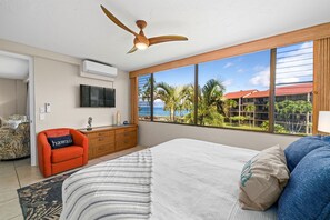 Une chambre avec un grand lit, un ventilateur de plafond, une télévision, une commode et une vue sur des palmiers et un immeuble.