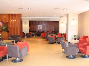 A hotel lobby with multiple seating areas, including red and grey chairs, round tables, and a wooden accent wall. - Magic Games (Oropesa del Mar)