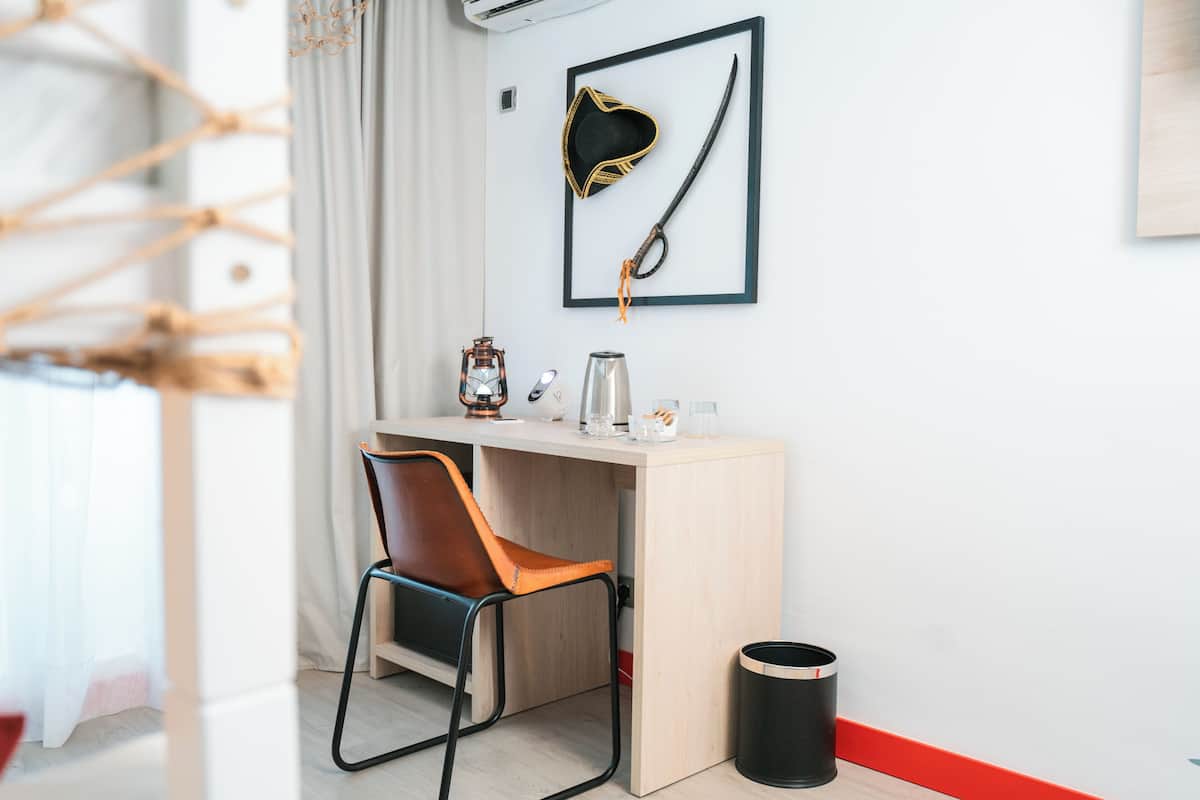 Une chambre moderne avec un bureau en bois, une chaise, un tableau encadré représentant un chapeau et un sabre de pirate, une machine à café et une poubelle.