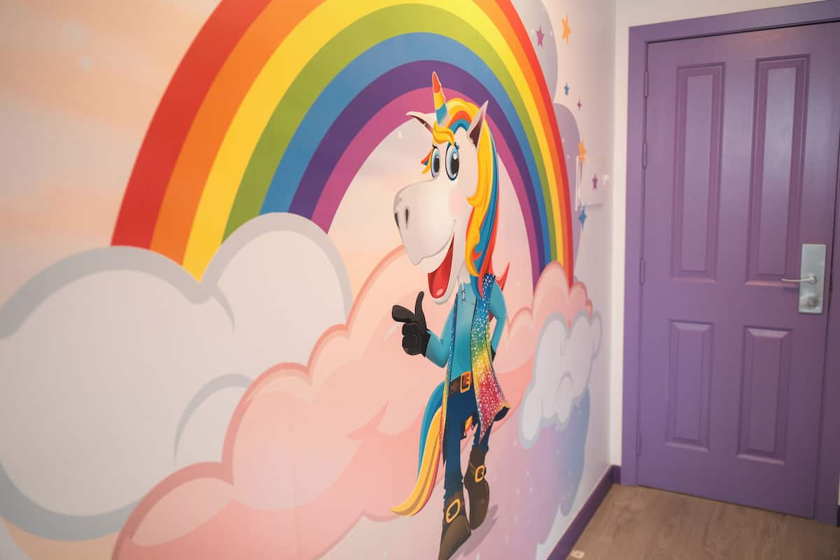Un mur orné d’une fresque représentant une licorne et un arc-en-ciel, une porte violette et un plancher en bois.