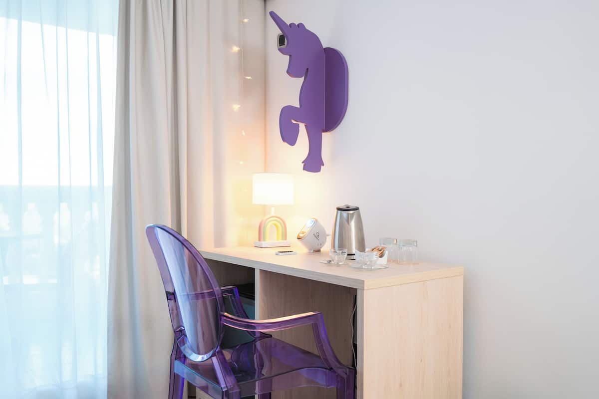 Une chambre d’hôtel équipée d’un bureau, d’une décoration murale représentant une licorne violette, d’une lampe, d’une machine à café et d’une chaise transparente.