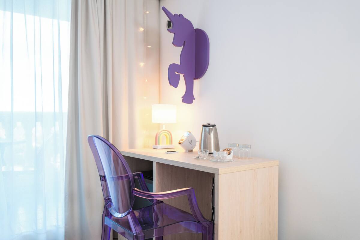 Ein Hotelzimmer mit Schreibtisch, einer Wanddekoration mit einem violetten Einhorn, einer Lampe, einer Kaffeemaschine und einem transparenten Stuhl.