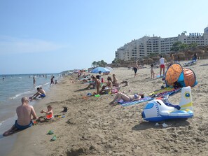 A beach with people sunbathing, swimming, and playing. There are umbrellas, beach chairs, and a tent. A hotel is visible in the background. - Magic Fantasy (Oropesa del Mar)