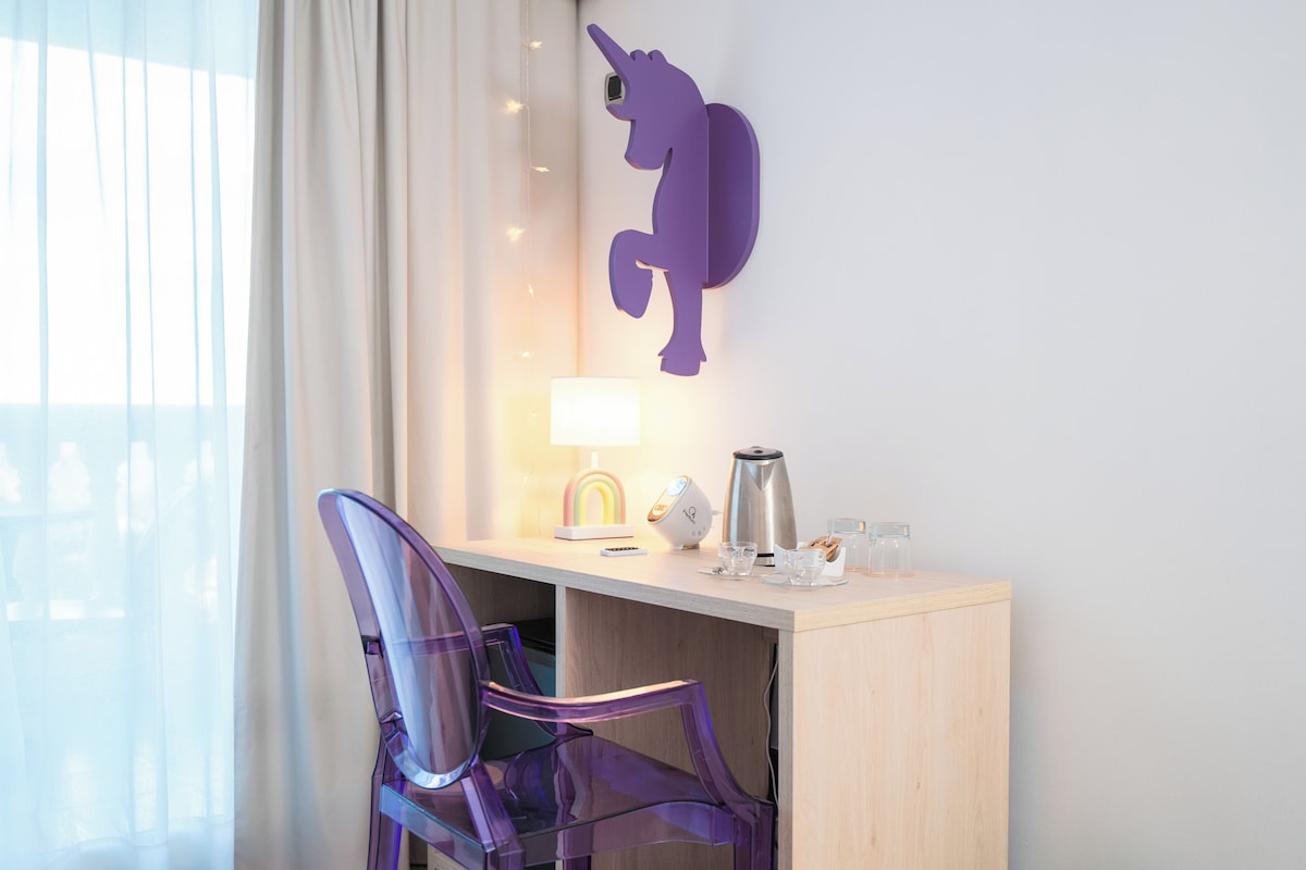 A hotel room with a desk, a purple unicorn wall decoration, a lamp, a coffee maker, and a clear chair.