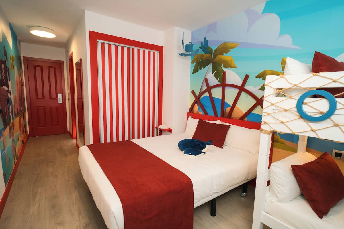 Une chambre d’hôtel avec un lit superposé, une porte à rayures rouges, une fresque représentant des palmiers et un mur à rayures rouges et blanches.