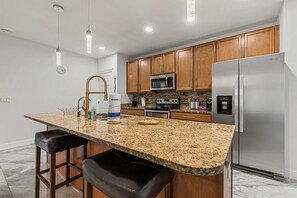 Une cuisine dotée de plans de travail en granit, d’armoires en bois, d’appareils électroménagers en acier inoxydable et de luminaires suspendus.