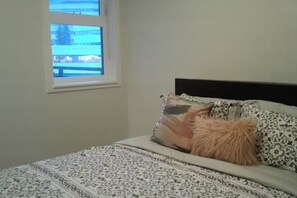 A bedroom with a bed, patterned bedding, a window with a view of buildings, and a headboard.