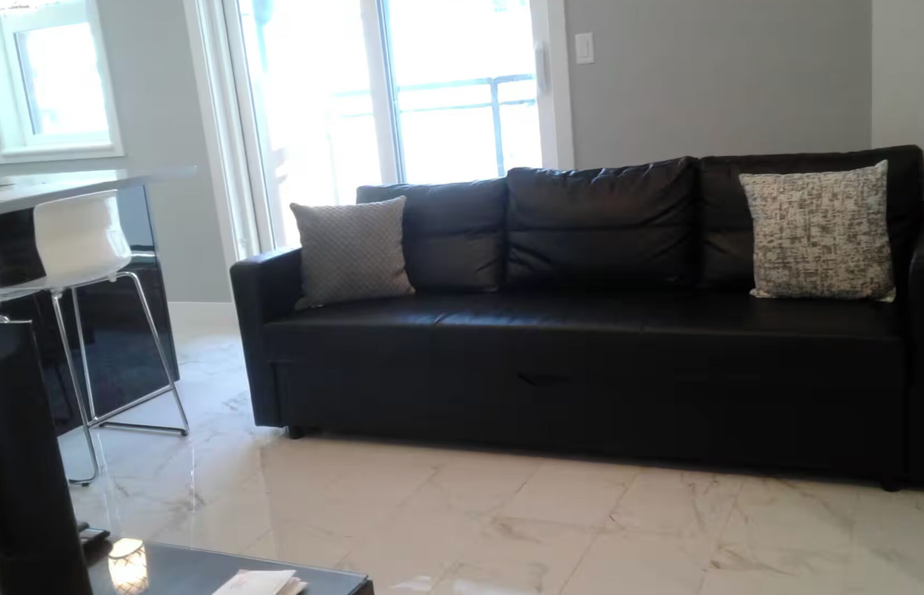 A black sofa with cushions, a white chair, and a small table with a lamp in a room with large windows.