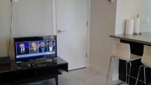 Ett rum med en tv som visar nyhetssändningar, ett skrivbord med en stol och en liten köksdel.