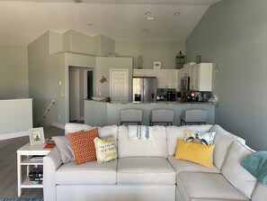 Ein modernes Wohnzimmer mit einem grauen Sofa, einem kleinen Couchtisch und einer Küchenzeile mit weißen Schränken und einem Edelstahlkühlschrank.