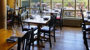 Un restaurante con mesas y sillas de madera, ventanas amplias con vista a un lago y vegetación.