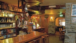 A bar area with shelves of bottles, a wooden counter, and stone walls.