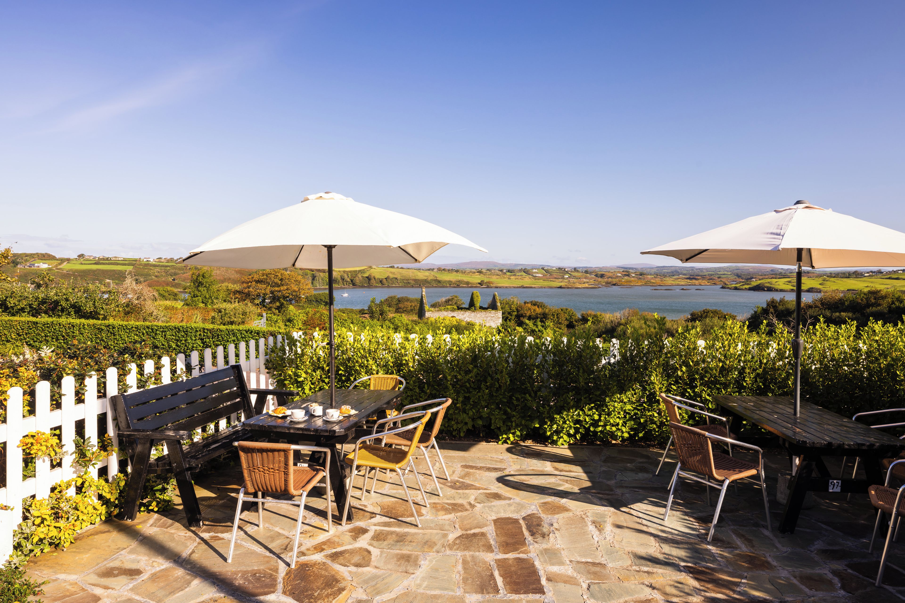 Eine Terrasse im Freien mit Steinboden, einem weißen Zierzaun und Blick auf einen See.