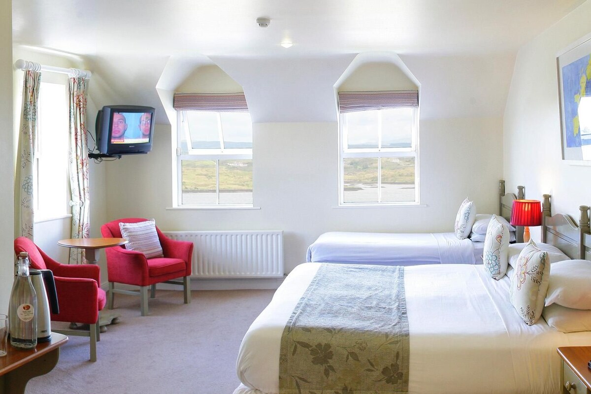 Quarto de hotel com duas camas, televisão, uma cadeira vermelha e vista para o campo.