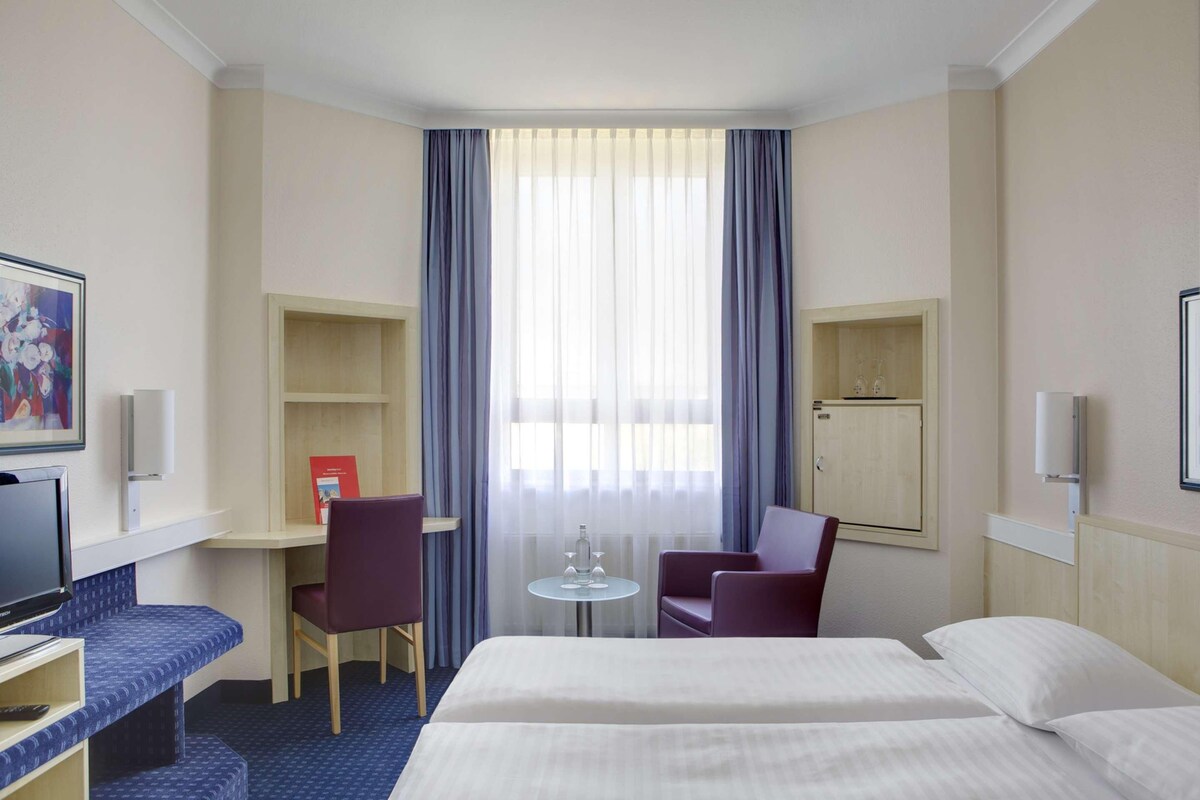 Habitación de hotel con cama, escritorio, silla, televisor y ventana con cortinas.