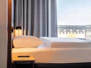 A hotel room with a bed, a nightstand, a lamp, and a view of buildings through the window. - B&B Hotel Kassel-wilhelmshöhe (Kassel)