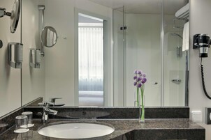 A modern bathroom with a granite countertop, a white sink, a mirror, a handheld hair dryer, and a glass shower enclosure.
