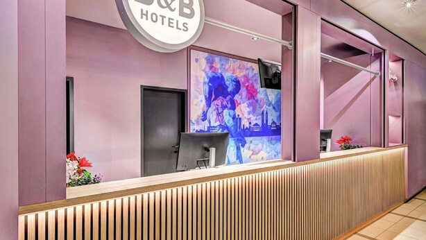 Ein Empfangstresen mit einem 'B&B Hotels'-Schild, einem digitalen Bildschirm und einer Holzpaneelen-Verkleidung.
