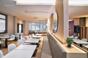 Un área de comedor moderna con mesas y sillas blancas, un largo banco y una partición de madera con una planta.