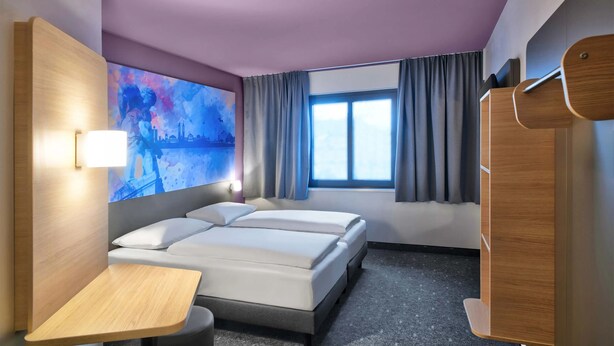A hotel room with two beds, a desk, a large window with curtains, and a wall-mounted TV. - B&B Hotel Kassel-wilhelmshöhe (Kassel)