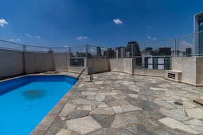 Une piscine sur le toit, avec un revêtement de sol en pierre et une clôture en métal, entourée d’immeubles de grande hauteur.