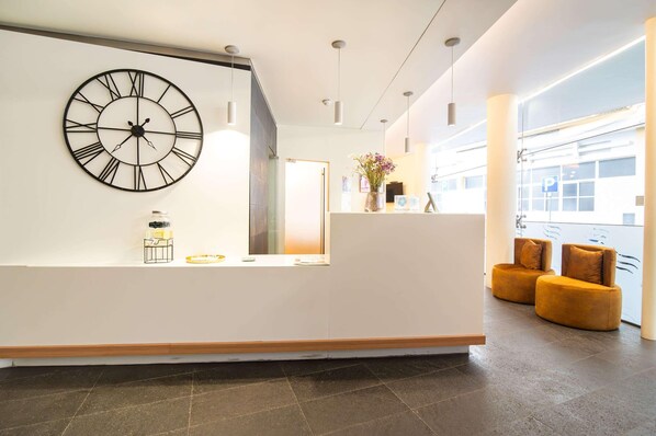 Un área de recepción con un gran reloj de pared, un mostrador de recepción, dos sillones acolchados y una ventana con vistas.