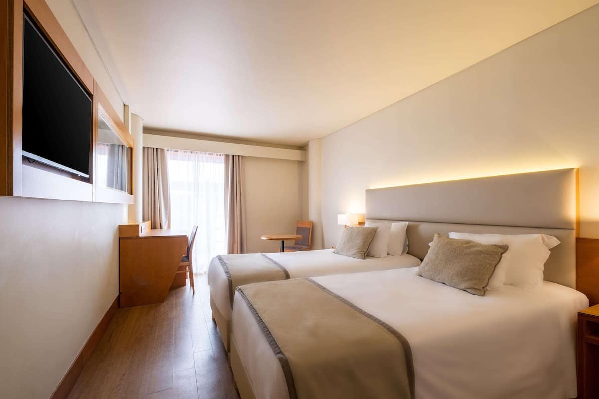A hotel room with two beds, a desk, a TV, and a window with curtains.