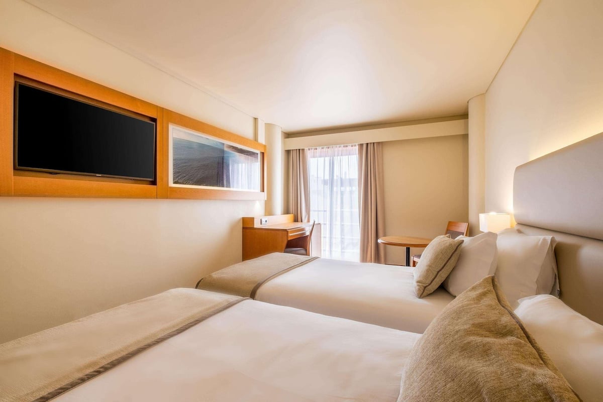 A hotel room with two beds, a desk, a chair, a TV, and a window with curtains.