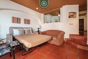 Un dormitorio con cama, sofá, una mesita y un espejo. Se ven unas escaleras que suben a otro nivel.