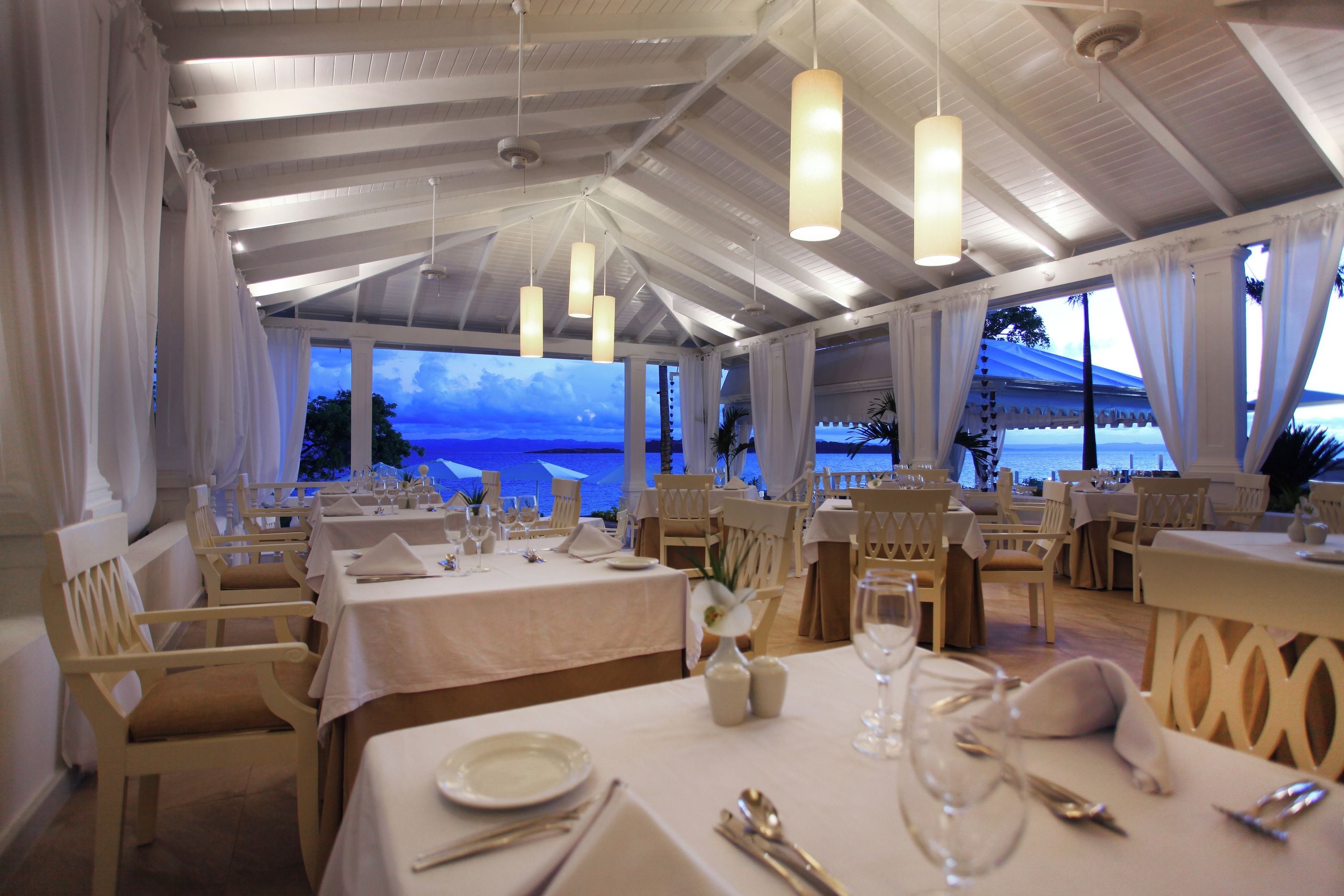 A restaurant with tables set for dining, white curtains, and hanging pendant lights.
