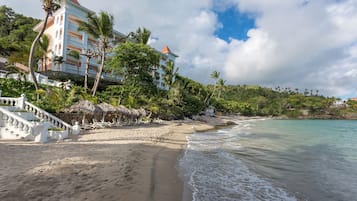 Un hôtel situé en bord de mer, avec un escalier menant à l’entrée, une plage dotée de parasols en chaume et une vue sur l’océan.