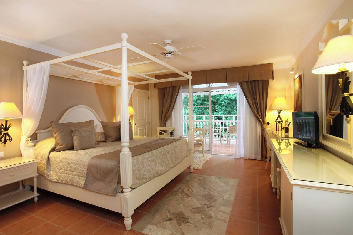 A four-poster bed with a canopy, a television on a dresser, and a balcony with a view of greenery.