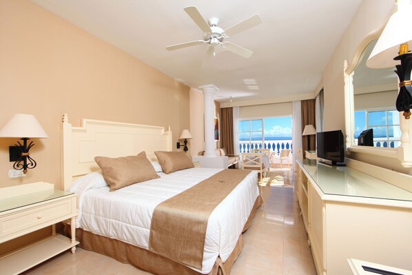 A hotel room with a bed, a TV, a ceiling fan, and a view of the sea.