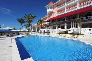 A swimming pool with lounge chairs and umbrellas, a multi-story building with balconies, and palm trees.