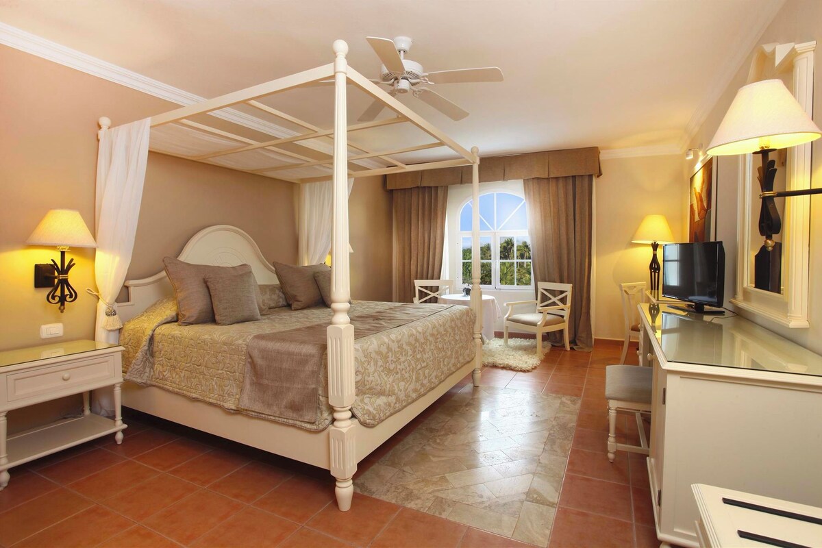 A four-poster bed with a ceiling fan, a television, and a desk with a chair in a well-lit room with a large window.