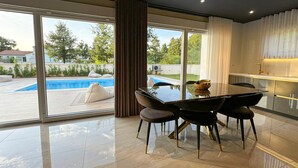 Ein moderner Essbereich mit einem Glastisch und Stühlen, Blick auf einen Pool sowie eine Küche mit weißer Arbeitsplatte.