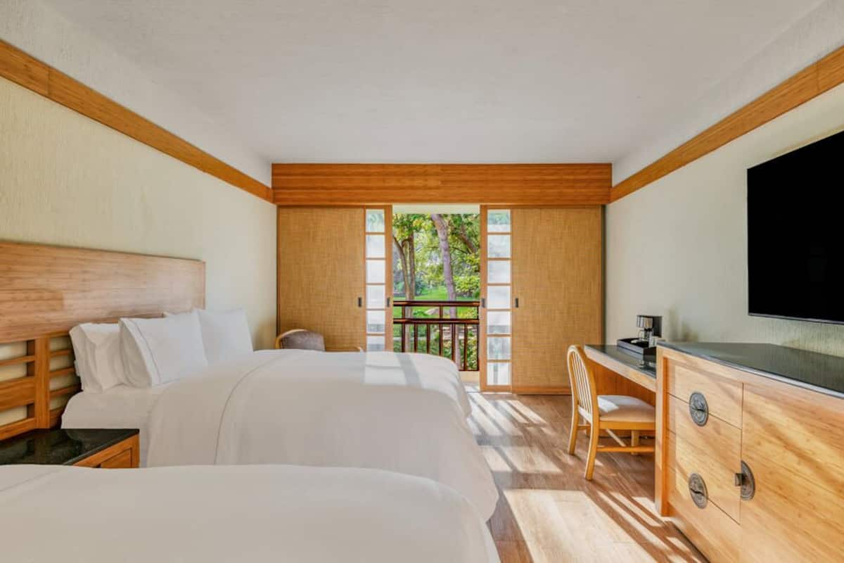 A hotel room with two beds, a desk, a TV, and a balcony with a view of greenery.