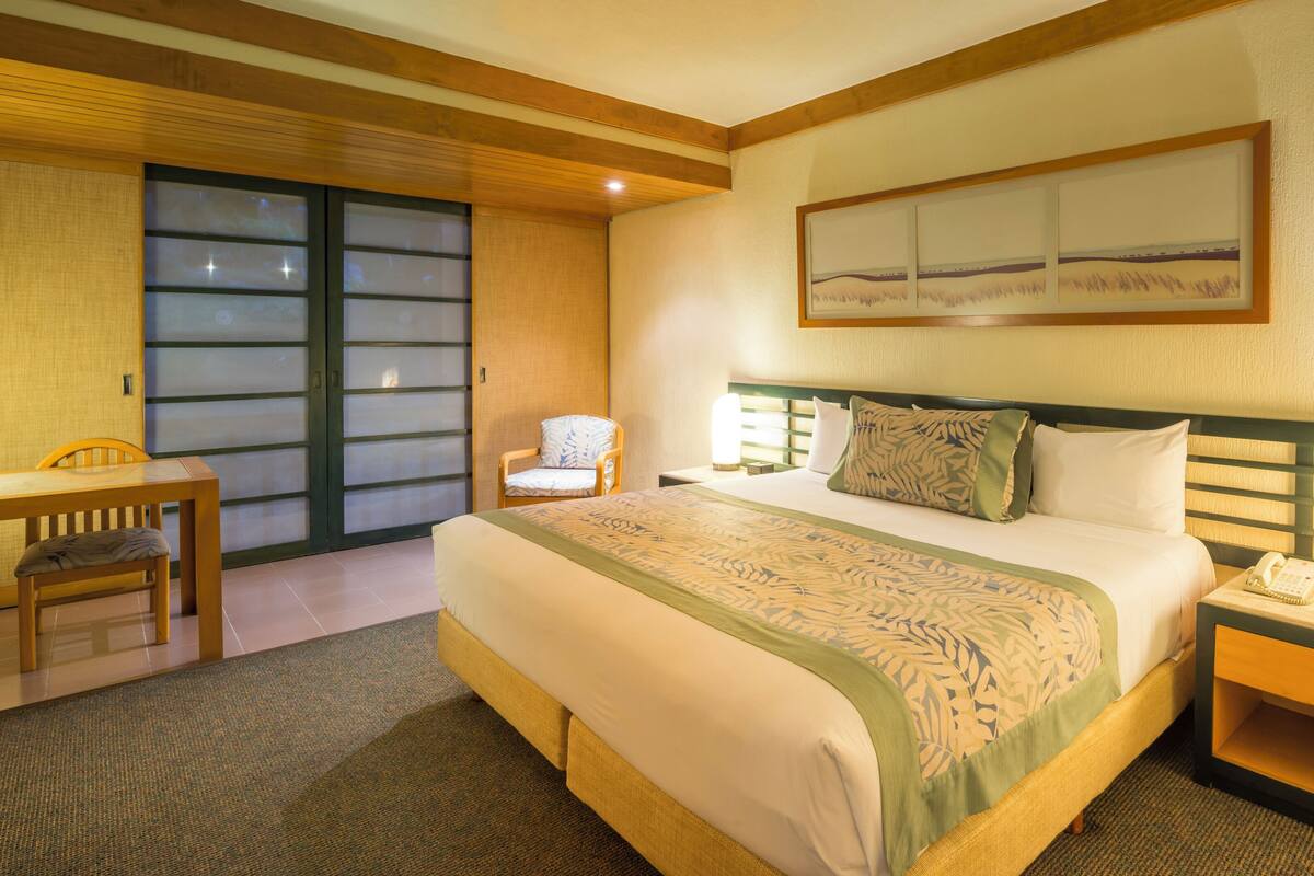 A hotel room with a bed, a chair, a desk, and a nightstand with a lamp.