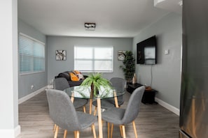 Un espace repas moderne avec une table en verre, des chaises grises, un canapé et un téléviseur fixé au mur.